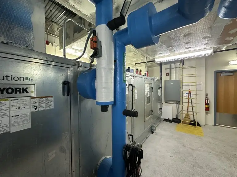 A commercial mechanical room featuring large blue insulated pipes connected to air handling units, with electrical panels and conduit visible in the background.