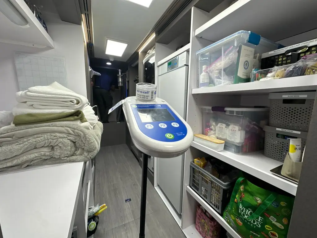Air quality monitoring device on a stand inside a narrow, well-organized laundry or utility room with shelves of supplies and folded towels.