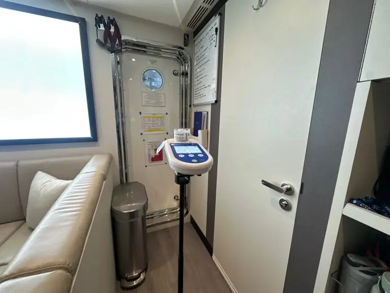 An Air-O-Cell air sampling device on a stand inside a yacht cabin, with a porthole, chrome piping, and a whiteboard visible in the background.