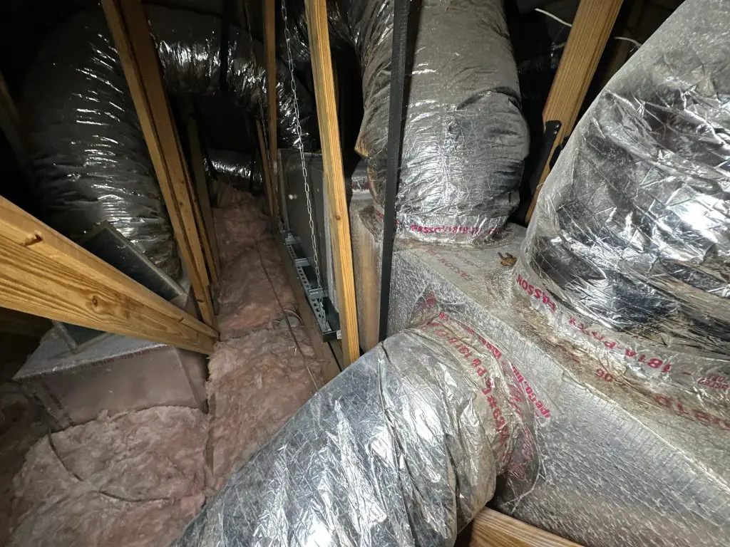 Attic space with insulated HVAC ductwork, wooden framing, and loose fiberglass insulation.
