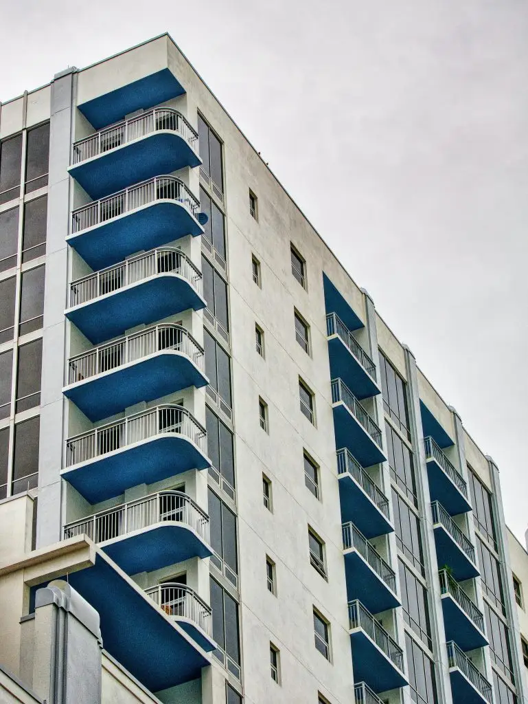 A close-up exterior view of a South Florida high-rise condominium building with rows of blue balconies and white stucco facade against an overcast sky.